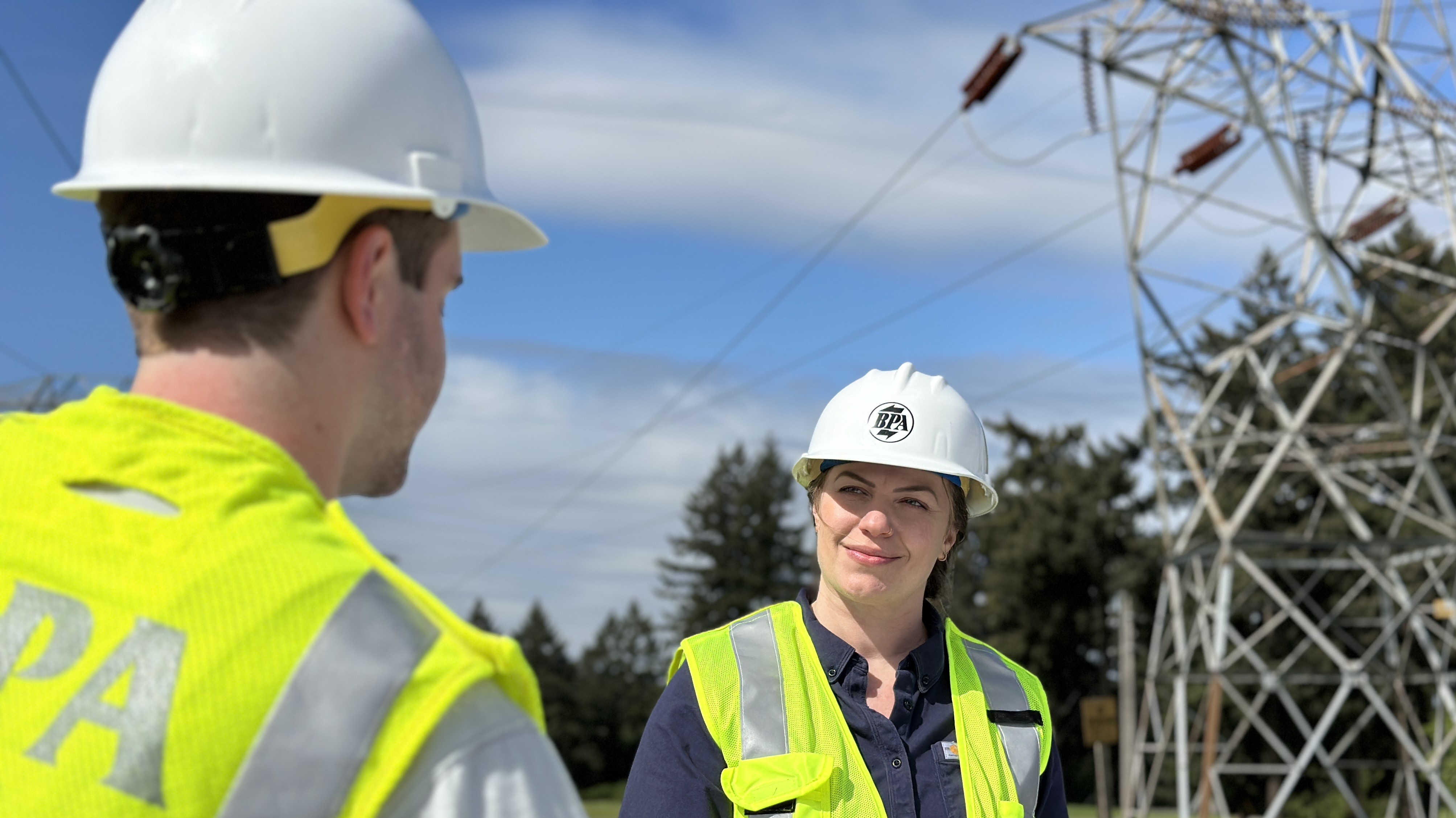 two people with white hard hats and neon safety vests, man with back to camera and woman looking toward man with transmission tower in background