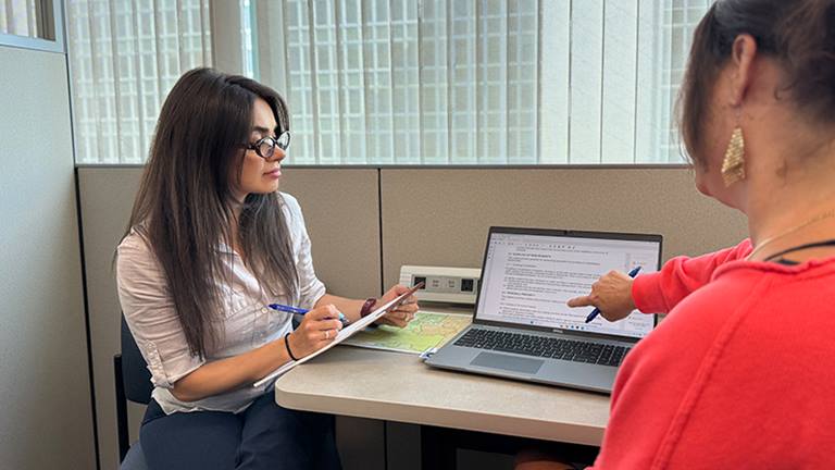 two women sitting at a desk looking at a laptop, while one woman points and the other holds a notepad and pen