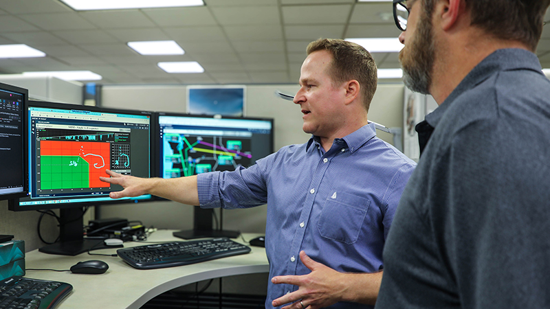 two men standing in front of a cluster of three monitors with one man pointing