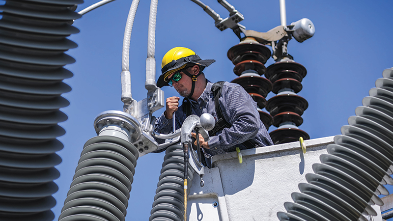 man in a bucket, wearing a long- sleeved shirt and hard hat, in the middle of large electrical metal structures