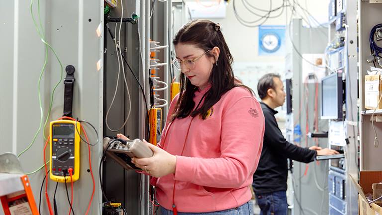 woman wearing glasses and pink sweatshirt, holding corded controller connected to a wall of wires with a man with dark hair in the background standing next to a similar wall looking at a monitor