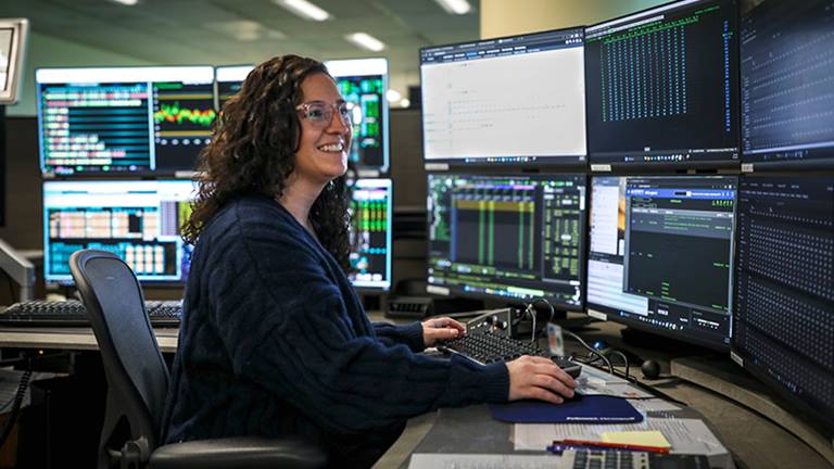 woman wearing a blue sweater and glasses, sitting in a chair at a desk with a hand on a keyboard and a hand on a mouse smiling at a cluster of six computer monitors