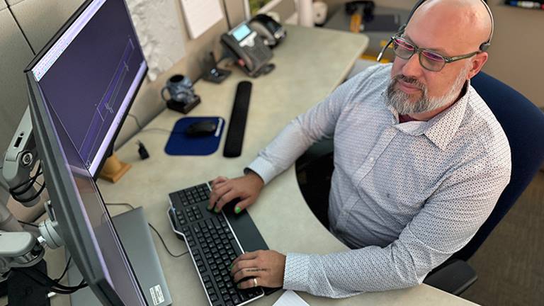 man wearing a long-sleeved shirt, glasses and a headset, sitting at a desk with his hands on a keyboard looking at two computer monitors