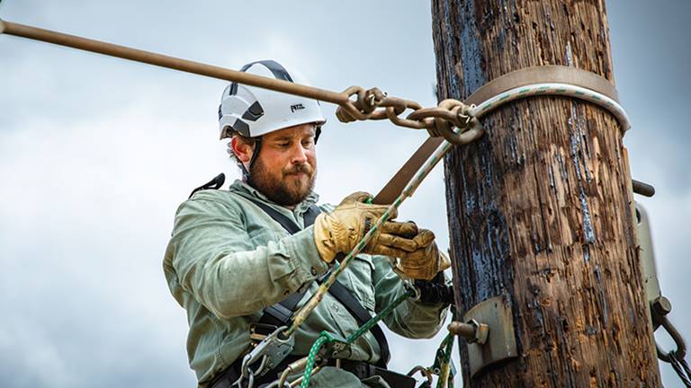 man wearing a long-sleeved shirt, gloves and a hardhat, attached to a harness attached to a wooden pole