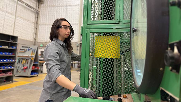 woman standing in front of large, caged equipment in a warehouse with her hand on a knob