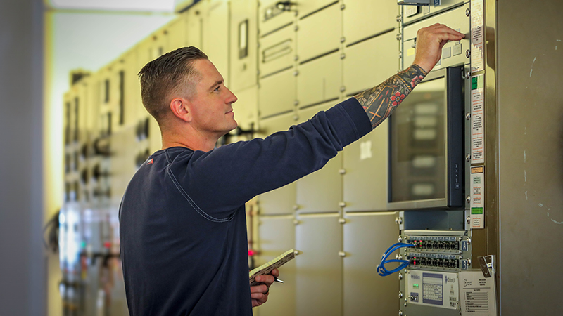 man holding a notepad in one hand with the other arm outstretched against a metal wall with various buttons and switches