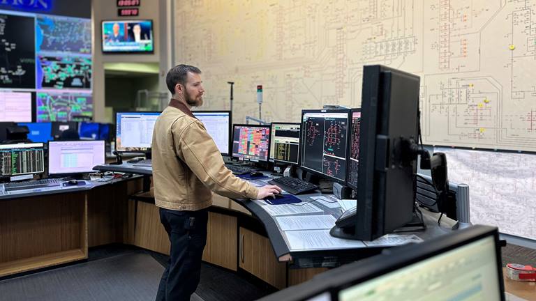 picture of man, standing in front of multiple computer monitors, in front of a wall with a lot of diagrams