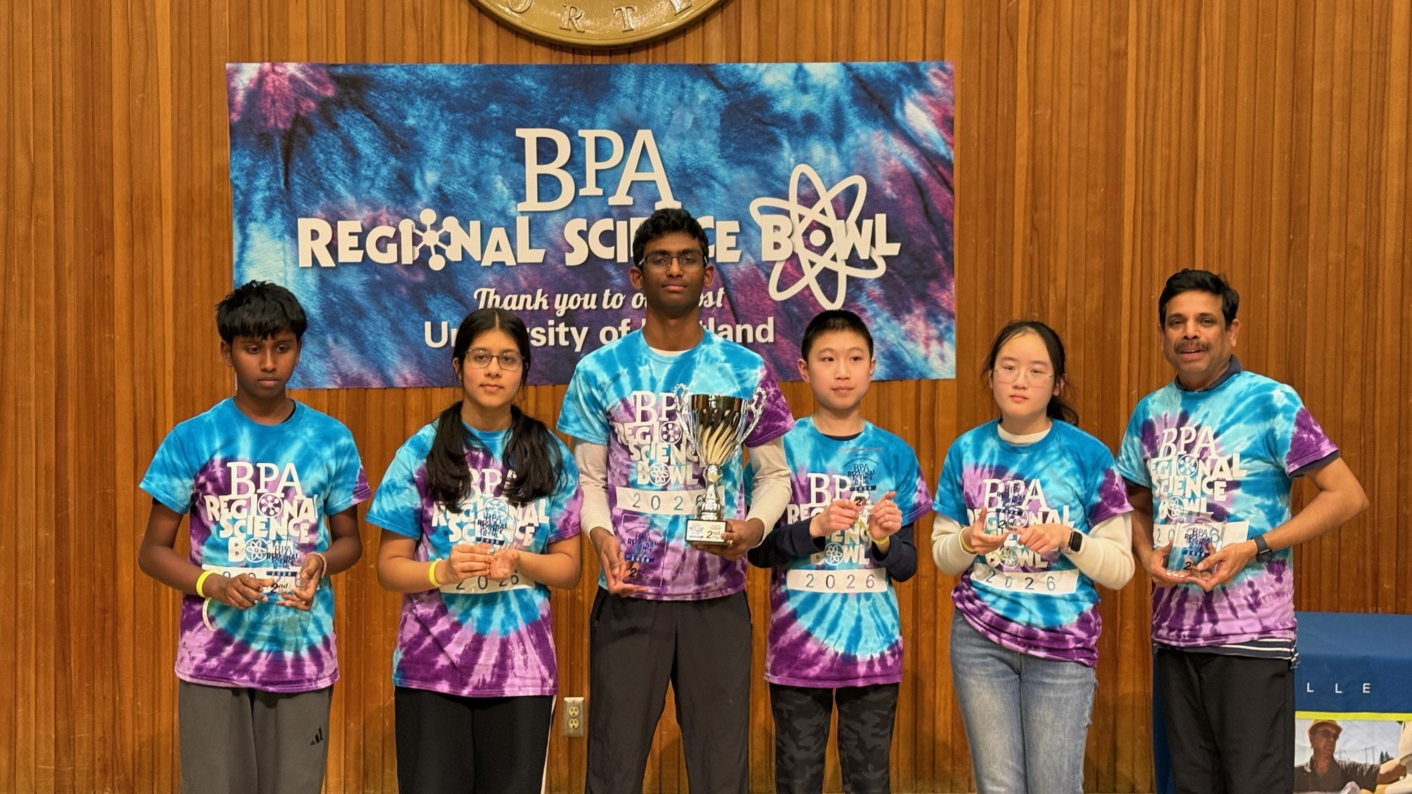 six individuals standing in front of a Science Bowl banner, holding trophies
