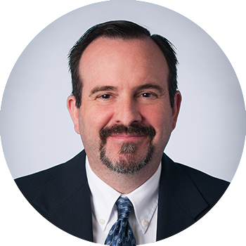 picture of man with dark brown hair, parted on the side, wearing a black suit with a white shirt and blue patterned tie, set against a grey background, photo in shape of circle