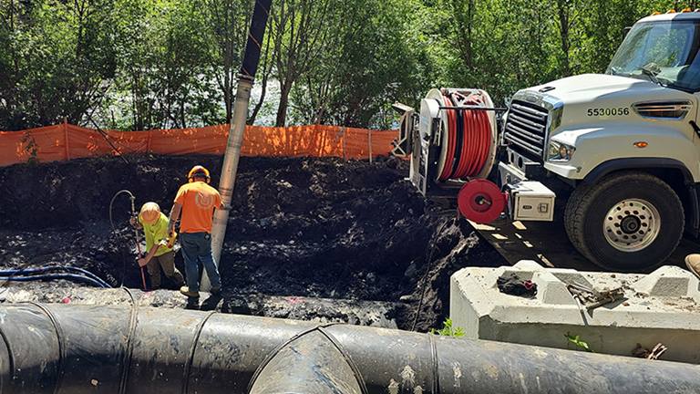 Large black pipe in foreground, behind which stands a man looking into a large hole while another man stands in the hole, near a truck, with the river in the background